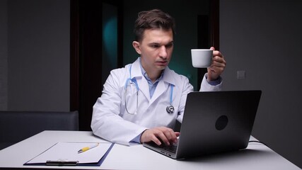 Male doctor in a white coat and stethoscope using a laptop for telemedicine consultations, working late hours, drinking coffee, and managing virtual healthcare at his desk