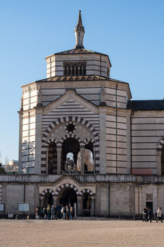 Octagonal striped tower and arched portico of Famedio chapel at Monumental Cemetery in Milan, Italy, with visitors gathered at entrance under clear blue sky.
