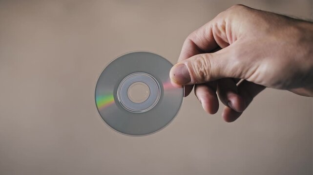 Top-down view of a male hand holding a small shiny compact disc. The rainbow iridescence on the surface highlights concepts of obsolete data storage and retro tech.