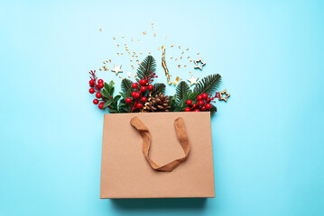 Craft paper bag with fir tree branches, red berries and pine cones on light blue background top view. Christmas festive shopping concept.