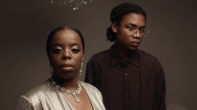 Portrait of Black woman wearing gem necklace with matching earrings posing next to Black man in deep brown designer shirt in fashion photoshoot standing against mocha toned wall in studio