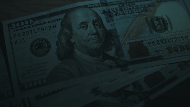 High angle close-up of a sweeping light illuminating stacked US one hundred dollar bills on a wooden surface. The dramatic low key lighting highlights Benjamin Franklin's portrait.