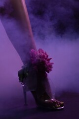 Woman wearing high heel with flower detail surrounded by purple haze at night