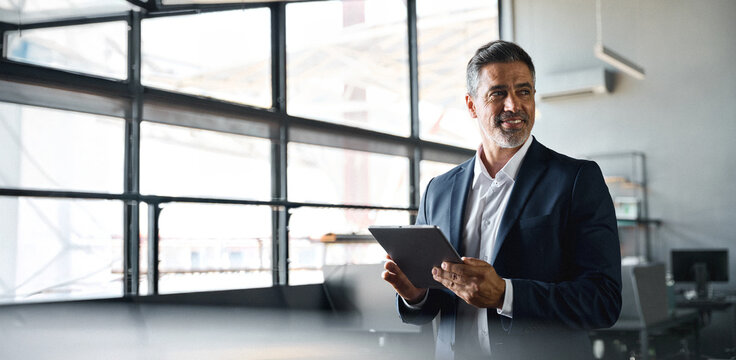 Happy middle aged business man ceo wearing suit standing in office using digital tablet. Smiling mature businessman professional executive manager looking away working on tech device. Banner.