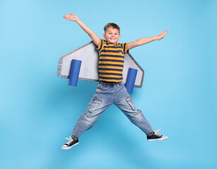 Little boy with cardboard plane wings jumping on light blue background