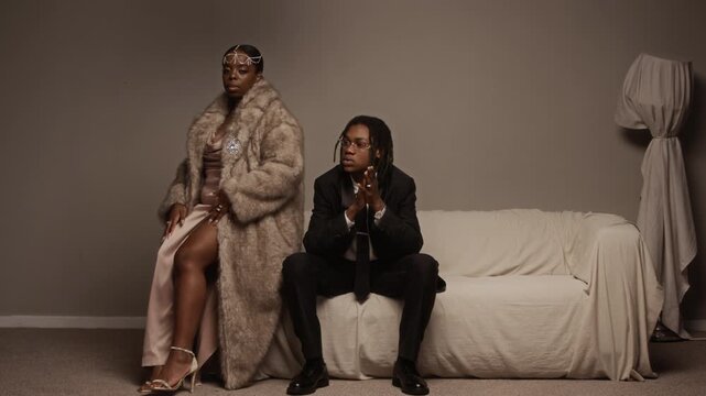 Full length shot of Black woman wearing fur coat over evening dress and Black man in black suit sitting on couch showcasing stylish jewelry during fashion photoshoot in mocha toned studio, copy space