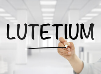 A conceptual image featuring the word lutetium handwritten in bold black letters on transparent glass, with a blurred modern white office interior in the background, representing this rare earth metal