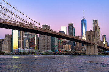 The iconic Brooklyn Bridge spans the East River, leading into the Lower Manhattan skyline during a peaceful purple dawn.