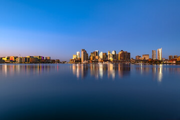 A peaceful panoramic view of the Boston skyline at dawn, showing the Seaport district, Downtown and West End reflecting on the still harbor water.
