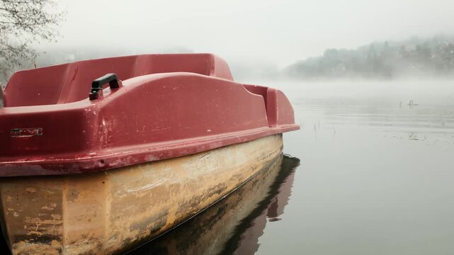 Abandoned pedal boat floating on a foggy lake with still water and soft mist. Moody outdoor scene showing neglect, silence and calm atmosphere in a quiet natural landscape. Horizontal 4k footage