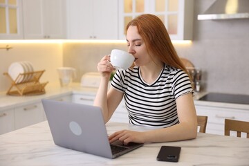 Woman having refreshing coffee while working on laptop at table in kitchen. Space for text