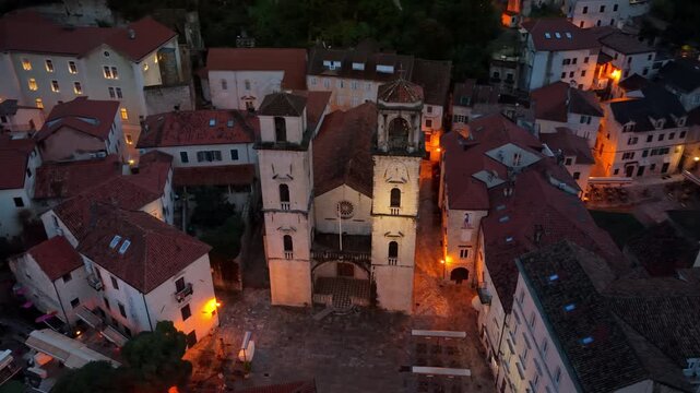 Aerial drone view of the Cathedral of Saint Tryphon surrounded by medieval stone buildings at dusk, showing the historic architecture in warm lighting. Kotor, Montenegro
