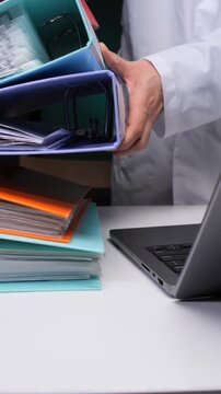 Vertical video healthcare professional organizing patient binders and digital files on a laptop, illustrating efficient medical records management and secure data handling