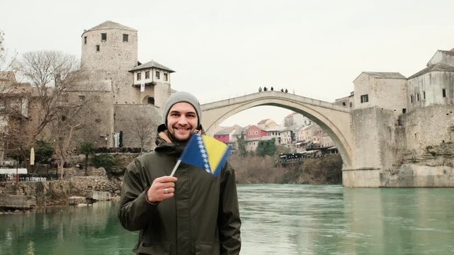 Young man waving the flag of Bosnia and Herzegovina near the historic Stari Most bridge in Mostar. Travel lifestyle scene with national identity, river landscape and iconic Balkan architecture