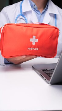 Vertical video doctor hands holding a bright red first aid kit with a white cross and first aid text, presenting it during an online consultation via laptop
