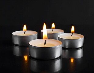 Four small, lighted candles with silver casings sit on a dark, reflective surface against a black background
