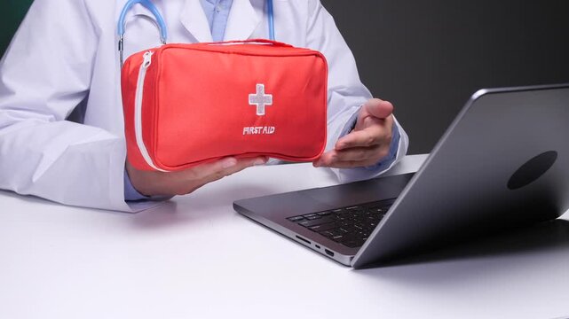 Doctor in lab coat discussing contents and use of an emergency first aid kit while sitting at desk with an open laptop, providing healthcare instructions online