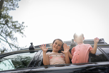 Siblings waving at camera from car sunroof on a sunny day. High quality photo