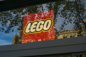 Fototapeta premium Barcelona, Spain, Europe - Nov 23, 2025: lego logo red yellow glass window storefront reflecting trees and historic architecture in a city street under blue sky with copy space for banner design