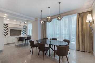 Fototapeta premium A dining area shows a round table with several chairs arranged around it. Natural light comes through large windows. The kitchen is visible in the background with cabinets.