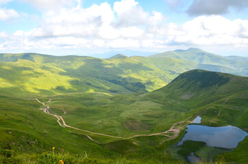 Lush green mountain valley features winding dirt paths across rolling hills toward a calm Dogyaska lake. Svydovets, Carpathian Mountains, Ukraine