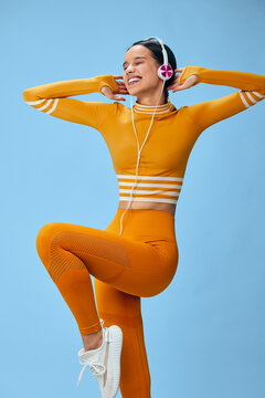 Woman in a bright orange fitness outfit dances with headphones on, capturing dynamic movement during a sport-inspired workout. Energetic pose against a blue backdrop highlighting strength, rhythm and