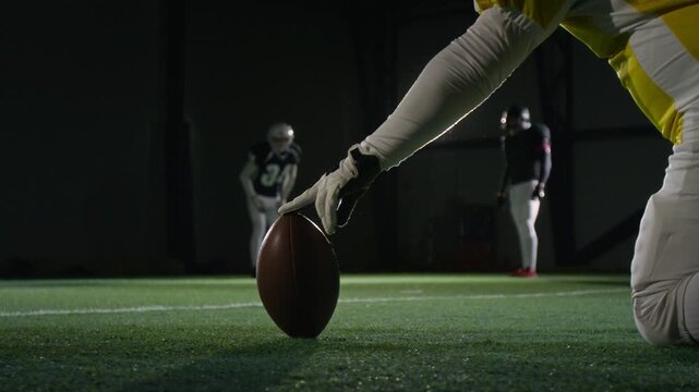 Medium ground level shot of sportsman holding ball and teammate practicing kickoff technique, while training with American football team at dark gridiron stadium, getting ready to play game