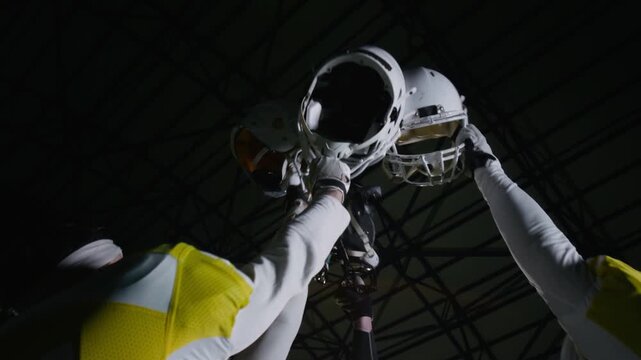 Medium tilt-up shot of four male athletes from amateur gridiron football team standing together at stadium, removing and lifting up helmets in display of unity while celebrating winning match