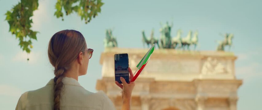 Woman recording a video of arch monument with her phone while holding Italian flag souvenir in Milan city