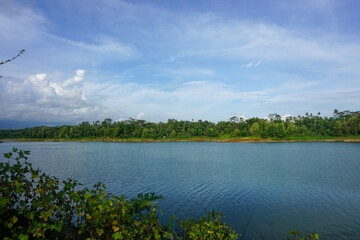 A lake with a clear sky above it. The water is calm and still. The trees surrounding the lake are lush and green, Malang, East Java, Indonesia.