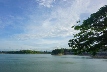 A lake with a clear sky above it. The water is calm and still. The trees surrounding the lake are lush and green, Malang, East Java, Indonesia.