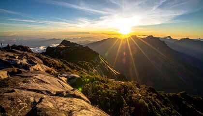 Mountain range sunrise golden sunburst over misty peaks, rocks, and sparse vegetation from an elevated foreground