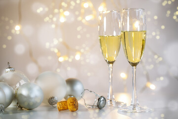 Two champagne flutes, Christmas baubles and the cork against a light warm background with some fairy lights and bokeh bubbles, festive holiday party like New Year, copy space