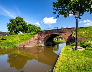 Quaint canal bridge with brick architecture reflecting in the calm water against a backdrop of lush green grass and blue sky
