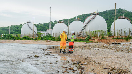 Two engineers, one in a yellow protective suit, test water on a beach. They use a laptop near large industrial gas tanks at a coastal plant facility.