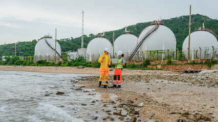 Two engineers, one in a yellow protective suit, inspect a coastal industrial site. They use a laptop and testing equipment on the beach near large gas storage tanks.