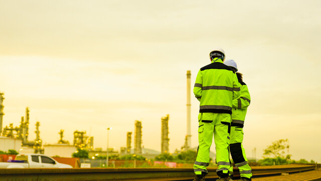 Two industrial workers in high visibility safety gear and hard hats stand on rusted metal pipes. They are inspecting a large factory or refinery plant during a golden sunset.