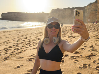 woman takes a selfie. A beautiful girl with a smartphone walks along the sea beach at sunset and takes a selfie. A girl smiles at her phone camera while standing on the beach at sunset.