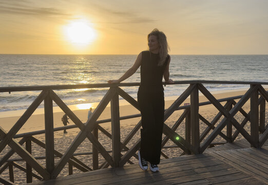 A wooden boardwalk overlooking the ocean. The boardwalk is empty and the sky is blue. The sun is setting and the water is calm