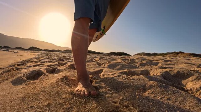 LENS FLARE, CLOSE UP: Bare feet of a male surfer walking along empty sandy beach in golden morning light and sand flying up with each step. Carefree surfing lifestyle on beautiful Canary Islands.