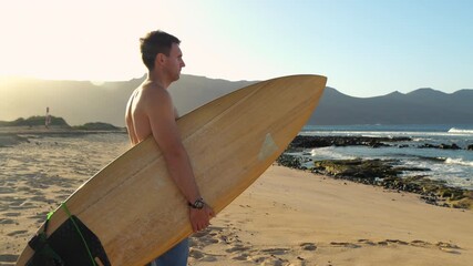 PORTRAIT, LENS FLARE: Man stands on beach, holding his surfboard while gazing at waves. Early morning light bathes the shoreline and volcanic landscape as he checks the waves before a surf session.