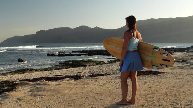 LENS FLARE, PORTRAIT: Woman is checking beautiful waves breaking in offshore winds on Lanzarote island. Surfer in skort and holding a wooden surfboard is getting ready for a morning surf session.