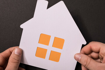 Close up shot of hands holding a paper house cutout with orange windows. Concept of homeownership,...