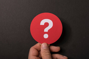 Close-up of a hand holding a red circle with white question mark against black background, symbolizing questions