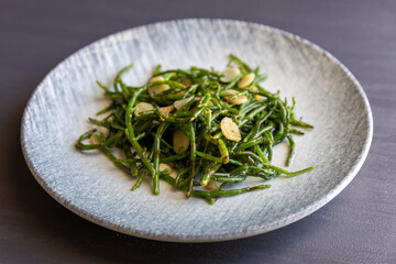 Fried green salty crisp samphire or salicornia plants with garlic on plate on grey background, close up