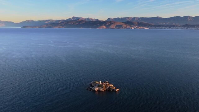 Aerial drone view of an isolated rocky Peristeres Island featuring the old lighthouse ruins, illuminated by golden hour sunlight against the deep blue Adriatic Sea. Greece