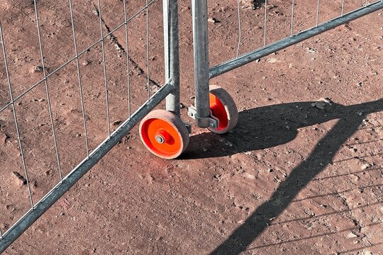 Orange wheels of a temporary metal fence casting long shadows on dusty ground, highlighting construction textures and urban infrastructure.