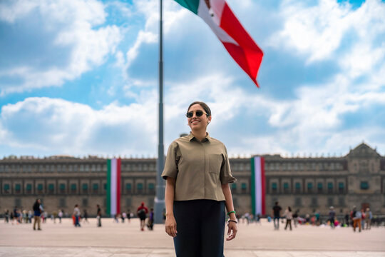 Smiling young Latin woman standing at Zocalo of Mexico City with the huge Mexican flag waving in the background under a bright blue sky, wearing casual clothes, sunglasses, showing national pride