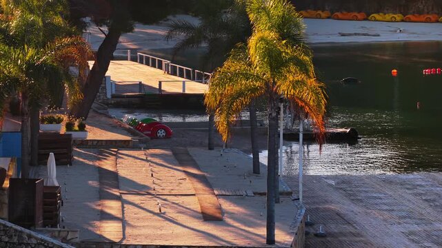 Aerial drone view of a beachfront area in Ksamil, with palm trees, colorful pedal boats, and calm water reflecting the warm morning light