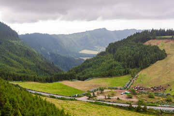 Green lush landscape of Sete Cidades, Azores, with a winding road and lake in the distance. Surrounded by mountains and dense forest. Located in Sao Miguel Island, Portugal.